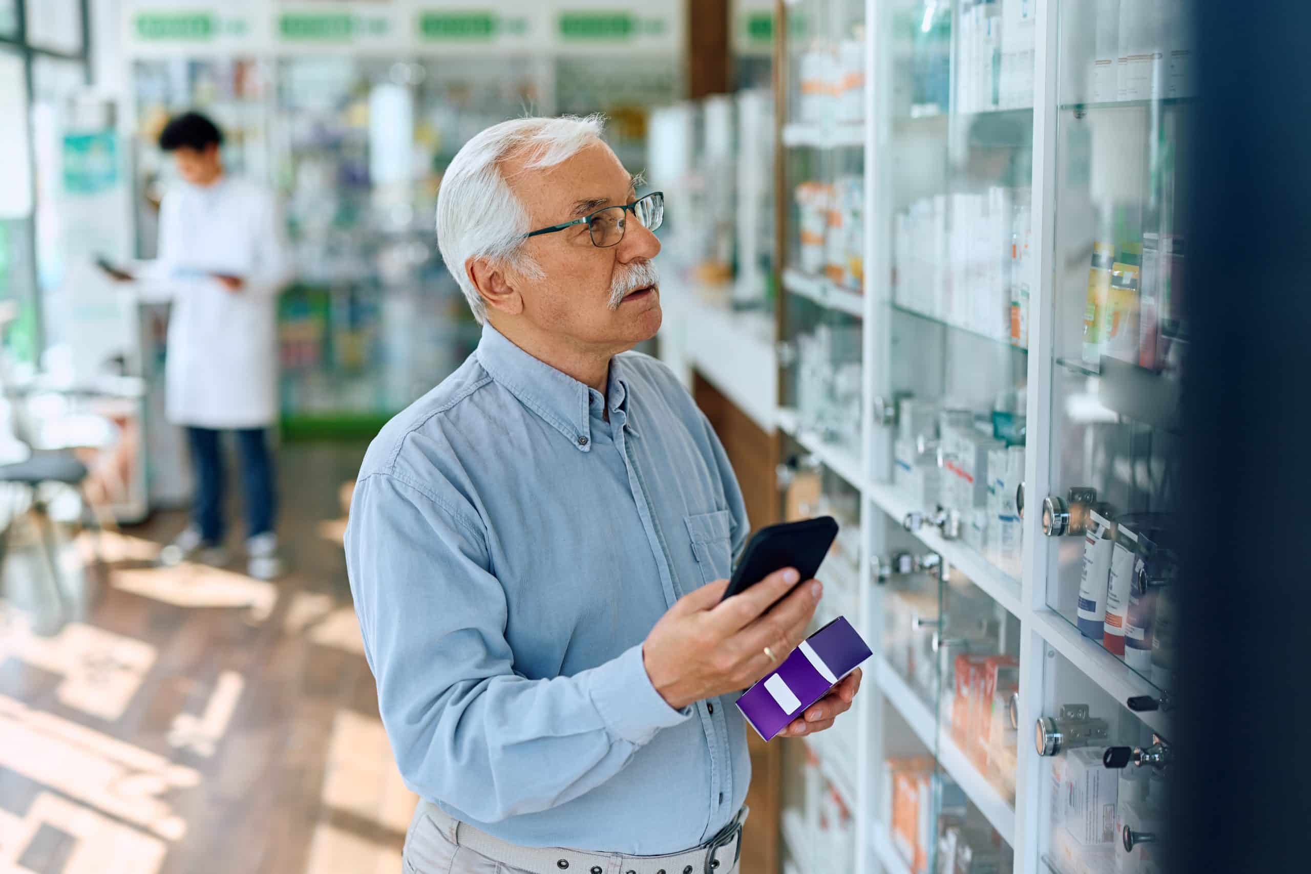 Senior man with glasses in a pharmacy, checking his phone while holding an OTC card, possibly researching who qualifies for OTC card benefits.